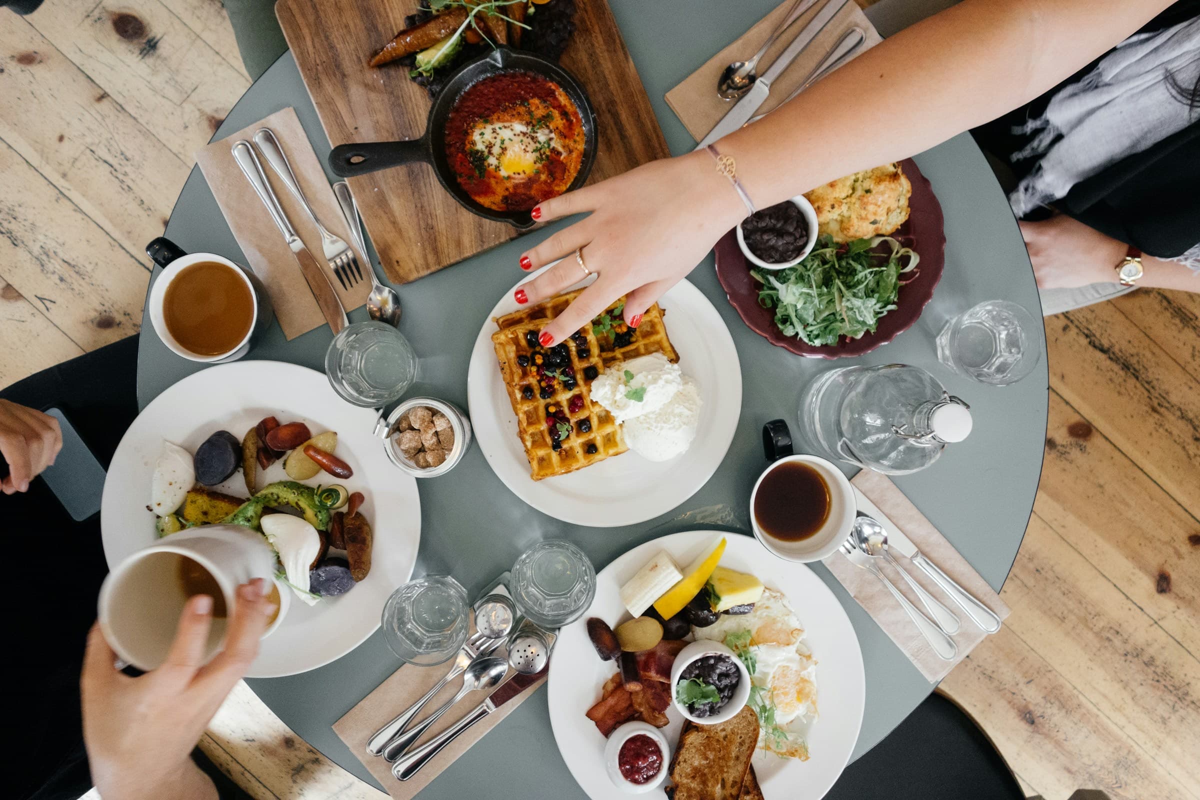 A brunch table seen from above, women's hands reaching for colorful plates